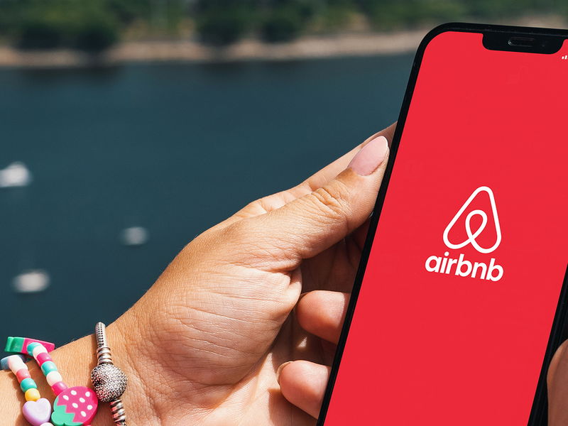 Girl holding smartphone with Airbnb app on screen. City and bay with some boats in the background. Rio de Janeiro, RJ, Brazil. March 2022.